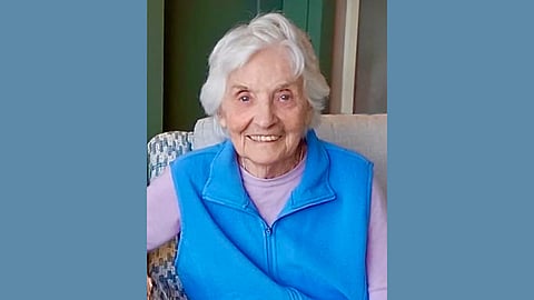 Photo of an older white woman wearing a lilac long sleeved shirt and bright cerulean blue zip up vest. She has white hair and a warm smile with kind eyes