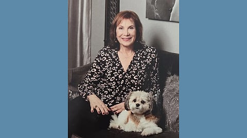 An older white woman with brown hair wearing a floral top sits smiling on a couch next to a small white and tan dog 