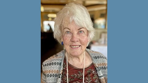 An elderly white woman with bright blue eyes smiles at the camera. She is wearing a burgundy floral top and a cardigan in fair isle print 
