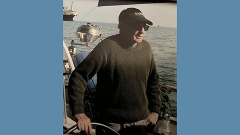 A middle aged white man stands at the wheel of a boat wearing a green sweater, sunglasses and a black baseball cap 