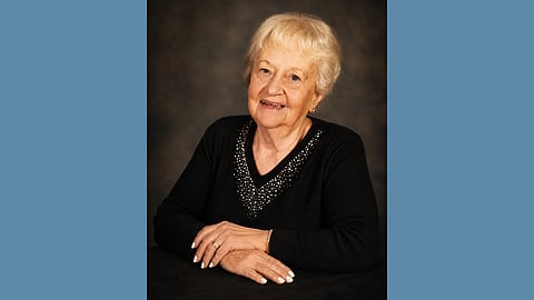 An elderdly white woman with white hair wears a black sweater with rhinestones around the v-neck. Her nails are painted and she's smiling 