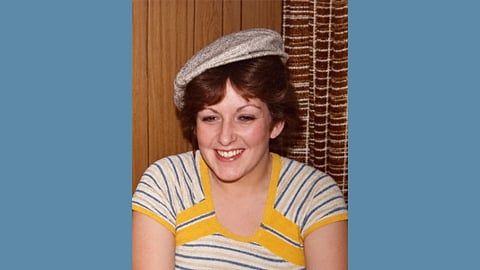 Vintage photo from the 1970s features a young brunette woman wearing a blue white and yellow striped top and a newsboy cap, smiling 