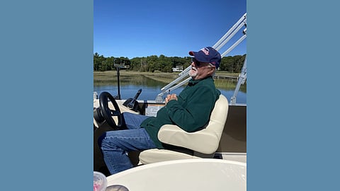 An older white man sits at the wheel of a boat wearing a hunter green jacket, sunglasses and a baseball cap