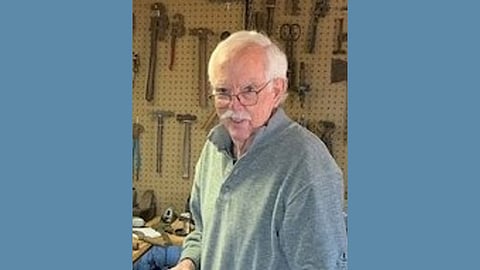 Older white man with white hair and mustache stands in a shop with tools behind him 