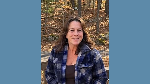Middle aged white woman with shoulder length wavy hair wears a blank tank top and blue checkered flannel shirt in a wooded area and smiles warmly at the camera 
