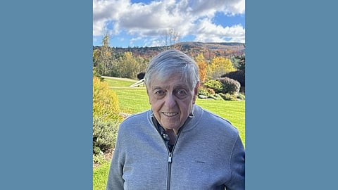 Elderly white man wearing a grey zip up jacket stands in a green valley with a beautiful blue sky behind him