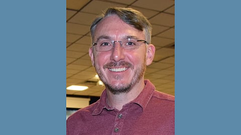 Middle aged white man wearing a rust colored polo and wire frame glasses. He has greying hair and a small beard. He is smiling warmly. 