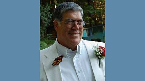 Older white man with grey hair and glasses wears a white tuxedo with a red rose on the lapel and a monarch butterfly on the other lapel 