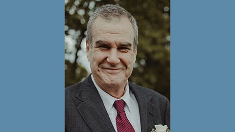 An older white main with silver hair wears a suit and tie and smiles at the camera with a closed mouth smile 
