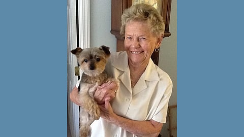Elderly white woman wearing a white button up shirt smiles at the camera as she hold a small black and tan dog 