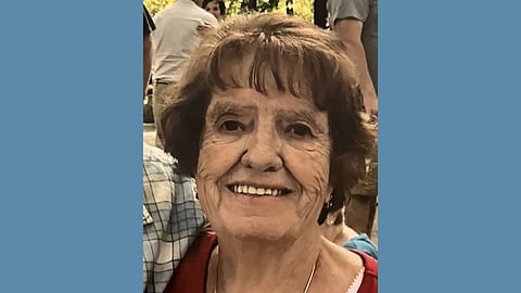 An elderly white woman with short brown hair wearing a red shirt smiles at the camera