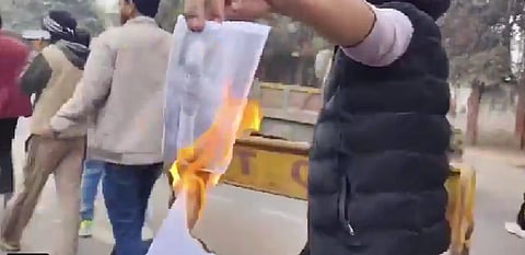  The youth holds a large printed image of Dr. Ambedkar aloft, lights it with a matchstick or lighter, and allows it to burn while surrounded by a small crowd of onlookers.