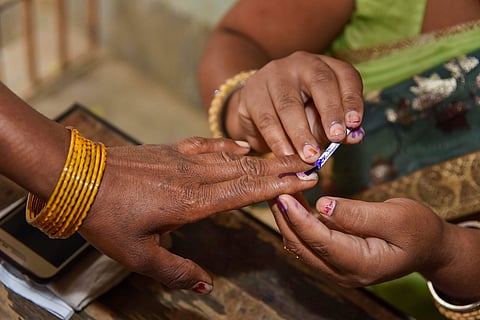 A close-up image showing a poll official applying indelible purple ink to the index finger of a voter's left hand during an election. The voter's hand is adorned with multiple yellow bangles. The official's fingers are also stained with ink.