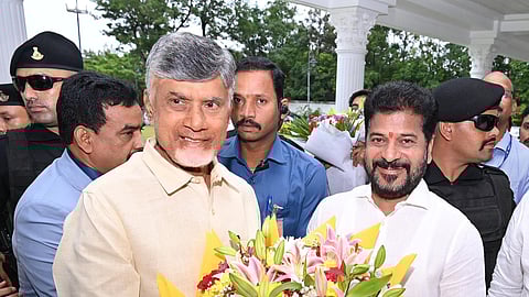A medium shot shows two men standing together, smiling and holding a large bouquet of colorful flowers. The man on the left, N. Chandrababu Naidu, has grey hair and a light beard, and is wearing a light yellow button-up shirt. The man on the right, Revanth Reddy, has dark hair and a beard, a red mark on his forehead, and is wearing a white button-up shirt. The bouquet between them is filled with pink lilies, red roses, and yellow and purple flowers, wrapped in yellow paper. Several other men, some in blue shirts and others in black security uniforms with sunglasses and berets, are visible in the background. The setting appears to be outdoors near a white building with pillars.