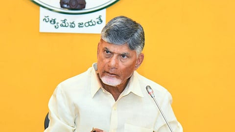 Andhra Pradesh Chief Minister Nara Chandrababu Naidu in a light yellow shirt sits at a desk, speaking into a microphone. He holds a pen and has documents in front of him. Behind him is the Andhra Pradesh government's emblem on a yellow wall.