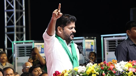 Telangana CM Revanth Reddy in a white shirt and green scarf speaks passionately at a podium adorned with colorful flowers. He raises one hand while addressing a crowd in an indoor setting with metal structures and people in the background.