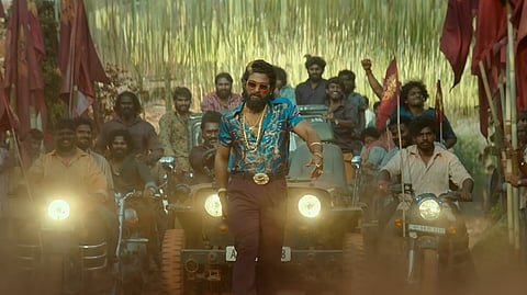 A frame from the film Pushpa 2: The Rule in which Allu Arjun wearing a blue patterned shirt and sunglasses walks confidently on a dusty path, flanked by motorcycles and jeeps. A group of people follows, cheering and raising hands. There are red flags in the background.
