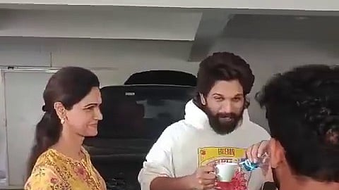 Allu Arjun in a white hoodie holding a cup of coffee. His wife Sneha Reddy is seen standing to his right in a yellow kurta. Both of them are smiling. 