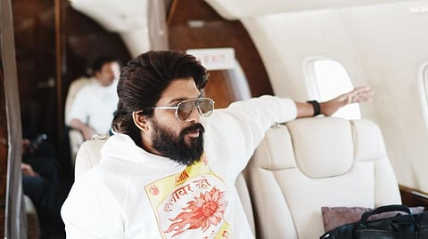 Allu Arjun in a white hoodie sitting inside an aircraft. His left arm is on the empty seat next to him, and there's a phone on the table in front of him. He is wearing sunglasses. 