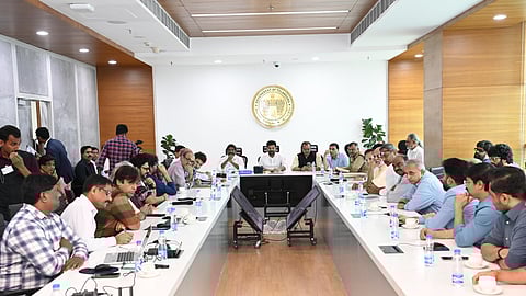 A  group of men are seated around a long conference table in a modern meeting room. Telangana Chief Minister Revanth Reddy is seated at the centre, surrounded by prominent Tollywood filmmakersand actors. There is a large emblem on the wall, and various individuals are engaged in discussion, some taking notes and others listening attentively.
