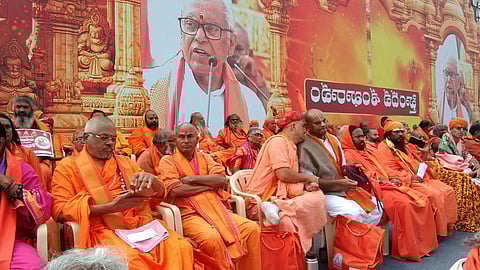 About 20 men in saffron robes are seating in two rows on a dais, with a poster that says 'Haindava Sankharavam' in Telugu, 