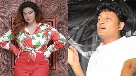 A woman in a floral shirt and red pants stands with hands on hips, next to a man speaking at a microphone.