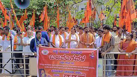 A group of people is gathered behind a banner, holding saffron flags. There is a large speaker on the left. Trees and a building are in the background.