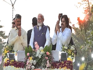 Chandrababu Naidu, PM Modi and Pawan Kalyan stand on a decorated platform outdoors, with flower arrangements in front of them. They are surrounded by greenery and a bright sky in the background.