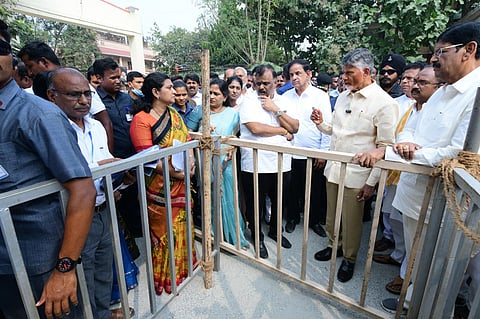 Andhra Pradesh Chief Minister Chandrababu Naidu in off-white shirt and trousers standing next to a barricade. With his left hand on the barricade, he is addressing officials and Ministers. He is surrounded by many men and women. 
