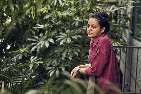 Payal Kapadia in a beige shirt standing on a balcony surrounded by a tree