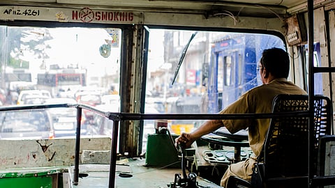 The driver of an Andhra Pradesh state transport bus in a brown shirt holding the gear road looking at traffic on the road ahead of him. The photograph is taken from behind, from a passenger's viewpoint. The road ahead is full of cars, buses and autos. 