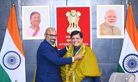 A formal photograph showing two individuals smiling warmly. BJP Nizamabad MP Dharmapuri is garlanding Union Minister Piyush Goyal with a turmeric garland while draping a golden shawl over his shoulders. Behind them are framed portraits of President Droupadi Murmu and Prime Minister Narendra Modi, the Indian national emblem, and the flags of India displayed on both sides