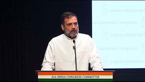 Rahul Gandhi iwearing a beard and a white kurta, speaking at a mic, set on a podium with a plaque saying All India Congress Committee