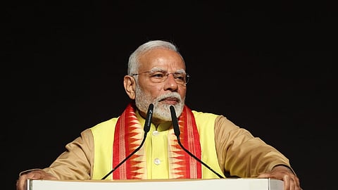 Prime Minister Narendra Modi standing at a podium with the emblem for Odisha Parba. He is wearing a brown shirt, yellow vest, and a white and red shawl. There are two mics in front of him. 