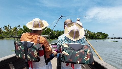 Two people wearing traditional attire and straw hats sit on a boat in Kerala's backwaters, with fishing nets and lush greenery in the background. 