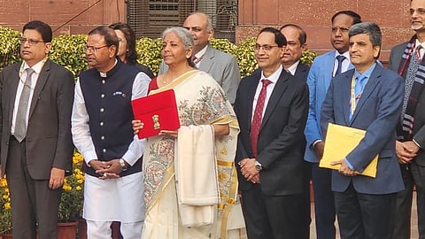 Finance Minister Nirmala Sitharaman and officials in front of the Ministry of Finance ahead of the budget speech. Minister wearing a white saree and red blouse. The budget documents are in a tablet carried in a red envelope held by the Minister. Officials around her are men in suits and one in kurta-pyjama. 