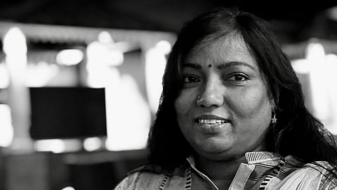 A black and white photograph of Sujatha Surepally wearing a striped collared shirt and a dupatta draped over her shoulder. She has long, dark hair, a small bindi on her forehead, a nose piercing, and earrings. She is smiling softly, and the background is blurred with lights and architectural elements visible.