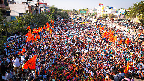 A large crowd of people gathered on a city street for a protest or rally. Many individuals are waving bright orange flags with symbols on them. The crowd includes men and women. Photographers and media personnel are capturing the event. The surrounding area consists of commercial buildings, trees, and vehicles.