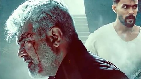 Actor Ajith in profile, facing right. He has grey hair and a beard and is wearing a dark-coloured jacket. The background is in different shades of grey.