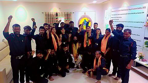 Men and women in black shirts, black pants, and a saffron scarf around their necks stand in a room facing the camera. One person sitting in the middle. Some of them have their fists raised as though raising a slogan. 