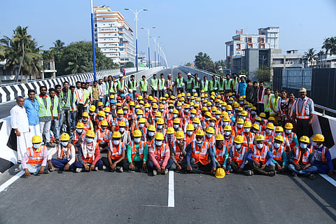 A large group of construction workers wearing hard hats and safety vests pose for a photo on a newly constructed road with buildings and greenery in the background.