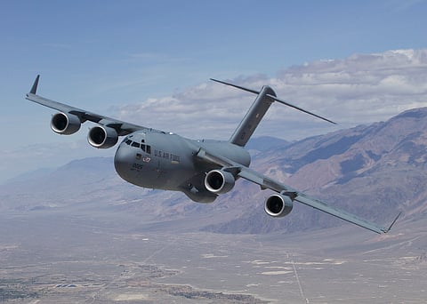 A grey US military plane in mid air, flying over hilly areas. The plane is barrelling towards its left. The words US Air Force can been seen near the cockpit windows. 