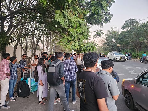 The terminated employees, some carrying backpacks and suitcases, stand on the side of a tree-lined road. Some individuals are engaged in conversation, while others appear to be waiting. 