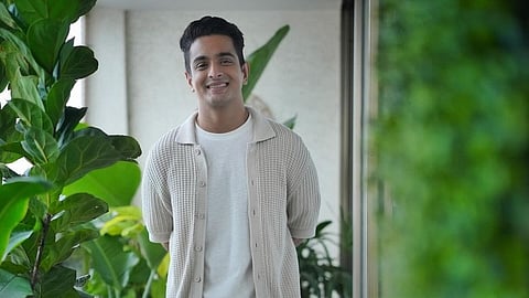 Ranveer Allahabadia with short black hair and a warm smile stands in a well-lit indoor space surrounded by lush green plants