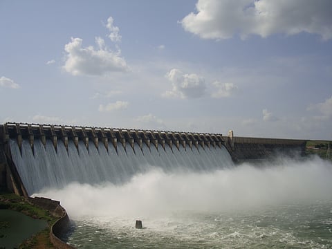 The image shows a large dam with multiple spillway gates open, releasing a massive flow of water. The structure appears to be made of concrete, and mist is rising from the water impact below. The sky is partly cloudy with blue patches visible. The surrounding landscape includes green vegetation and a river flowing downstream.