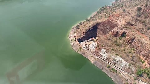 Aerial view of a body of water with a curved shoreline, where a rocky, arid landscape meets the greenish water. There is a construction or excavation site near the shore with visible machinery, materials, and a dirt road leading to it. Sparse vegetation covers the surrounding hilly terrain.