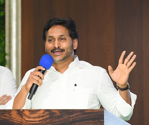 YS Jagan Mohan Reddy in a white shirt is speaking into a microphone with a blue foam cover while gesturing with his other hand. He stands behind a wooden podium, and a partially visible person in white is in the background. The setting has a dark wood-paneled wall.