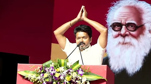 Actor and TVK president Vijay, wearing a white half-sleeved shirt, at a podium, with his hands folded in greeting above his head. The podium is red and decorated with purple, white and yellow flowers and leaves. The background is also red. There is a large picture of Dravidian icon Periyar behind him.