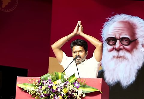 Actor and TVK president Vijay, wearing a white half-sleeved shirt, at a podium, with his hands folded in greeting above his head. The podium is red and decorated with purple, white and yellow flowers and leaves. The background is also red. There is a large picture of Dravidian icon Periyar behind him.