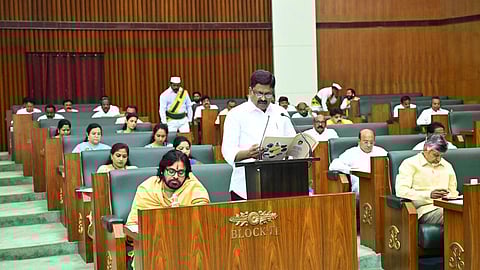 The image shows a legislative assembly setting with a man in a white shirt, AP Finance Minister Payyavula Keshav, standing at a podium, reading from a document. Several members are seated in green leather chairs arranged in rows, some listening attentively while others appear engaged in writing or reading. The setting has wooden paneling and an official atmosphere. There is a sign on the desk in front that reads "BLOCK-II." Some individuals are wearing traditional attire, and a few in the background are dressed in white with green and yellow sashes.